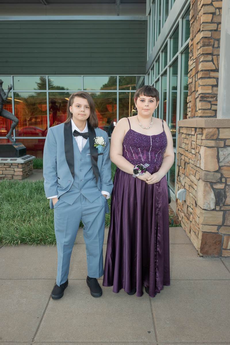 Two teens in formal wear posing outside a modern building: a boy in a light blue suit with a bow tie and corsage, and a girl in a purple gown with jewelry and a corsage.