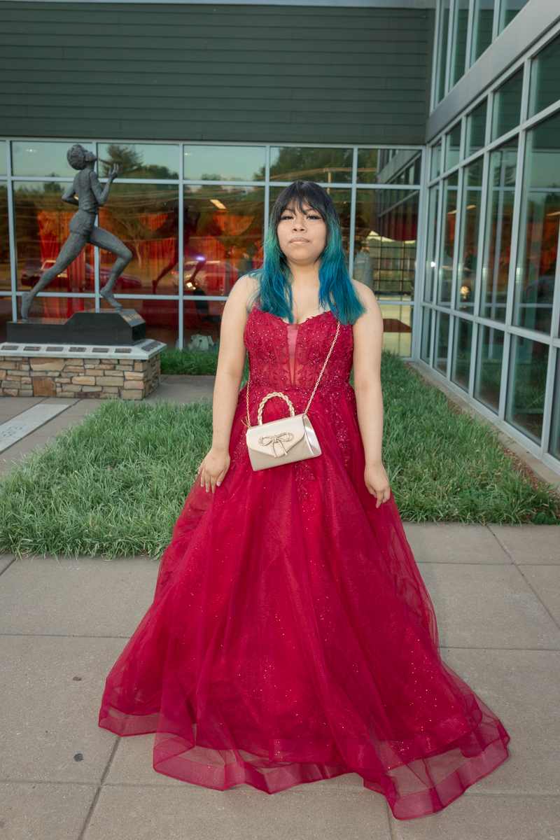Young woman in a red ball gown with teal hair standing outside a modern building.