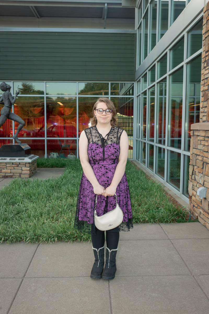 Young woman in a purple lace dress and black boots stands on a sidewalk outside a glass-fronted building, holding a white purse.