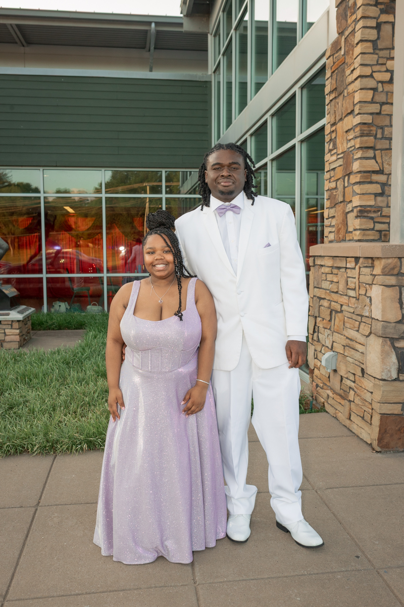 Couple in formal wear posing outside a glass-fronted building; woman in lavender gown, man in white suit with lavender bow tie.