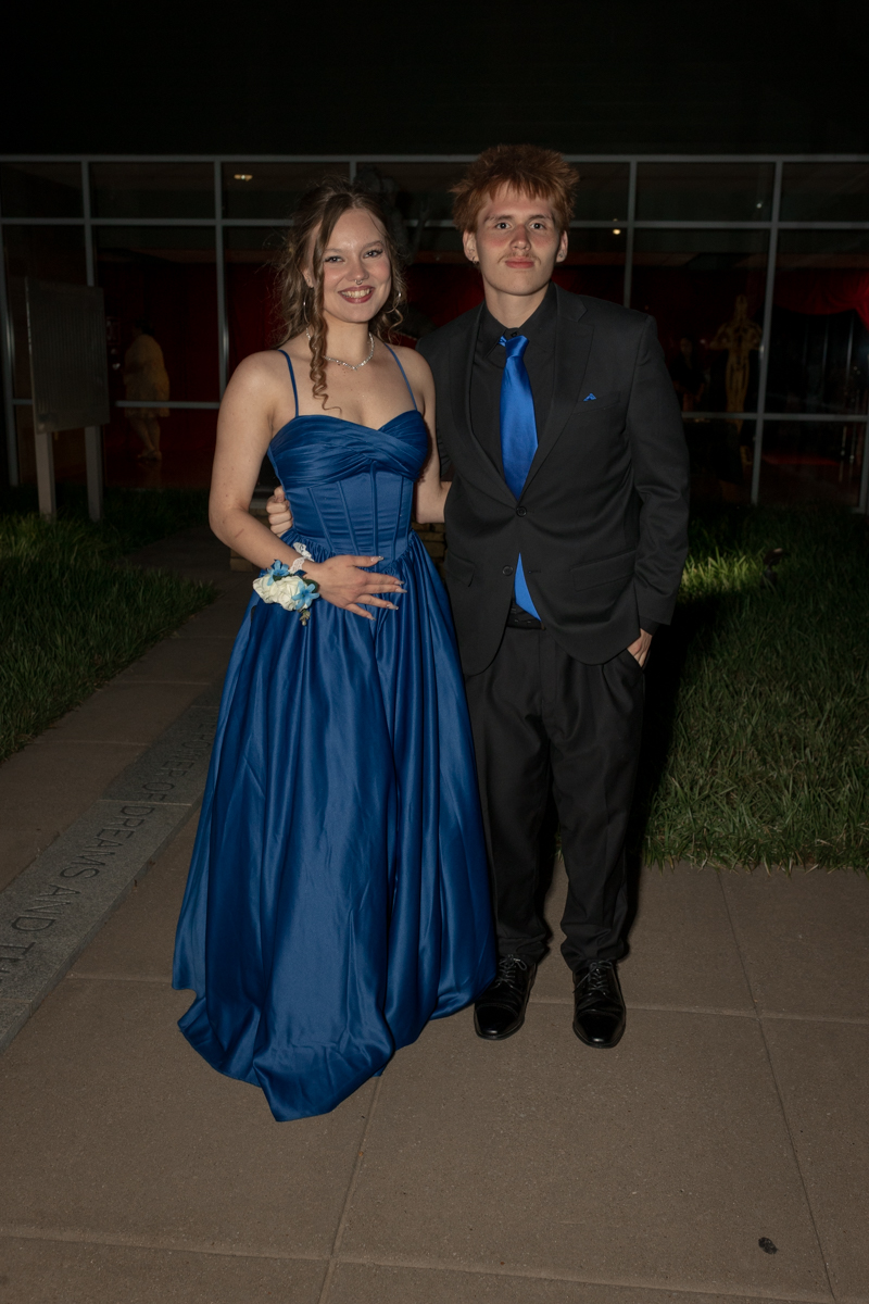 Two teens in formalwear pose together outside a venue; girl in a royal blue ball gown with a corsage and the boy in a black suit with a blue tie and pocket square.