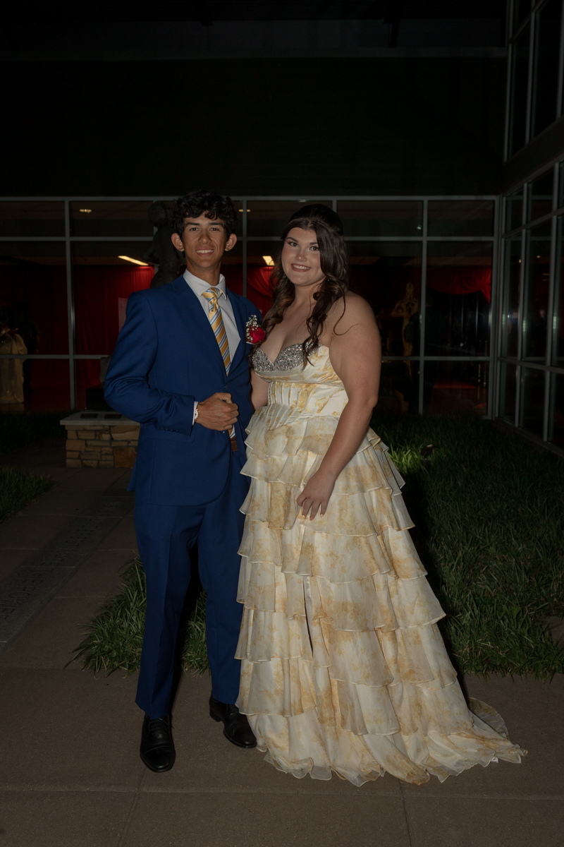 Young couple dressed for prom pose together outdoors; man in a blue suit with striped tie and boutonniere, woman in a tiered cream gown with silver bodice detail.