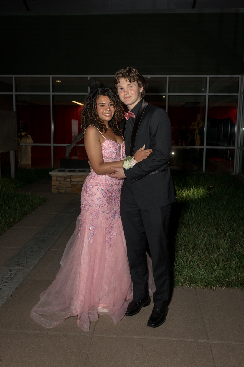 Young couple in formal attire posing together outside a building at night; girl in a pink sequined gown, boy in a black suit with a pink bow tie.