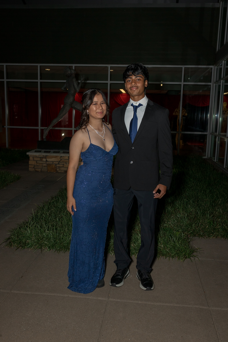 A couple dressed formally posing outside at night; woman in a blue sequined gown and necklace, man in a dark suit with a blue tie, in front of glass doors.