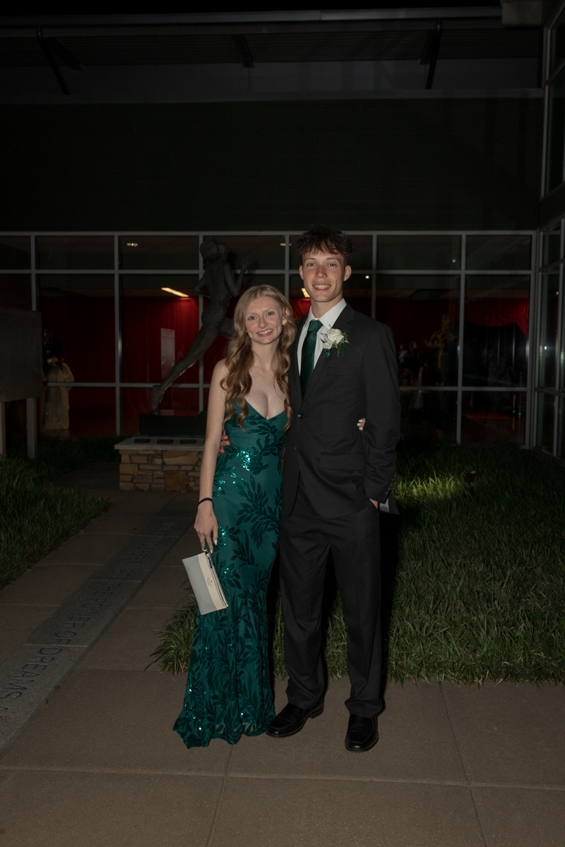 Young couple posing outside at night: woman in a teal sequined gown and man in a black suit with a white boutonniere, smiling together.