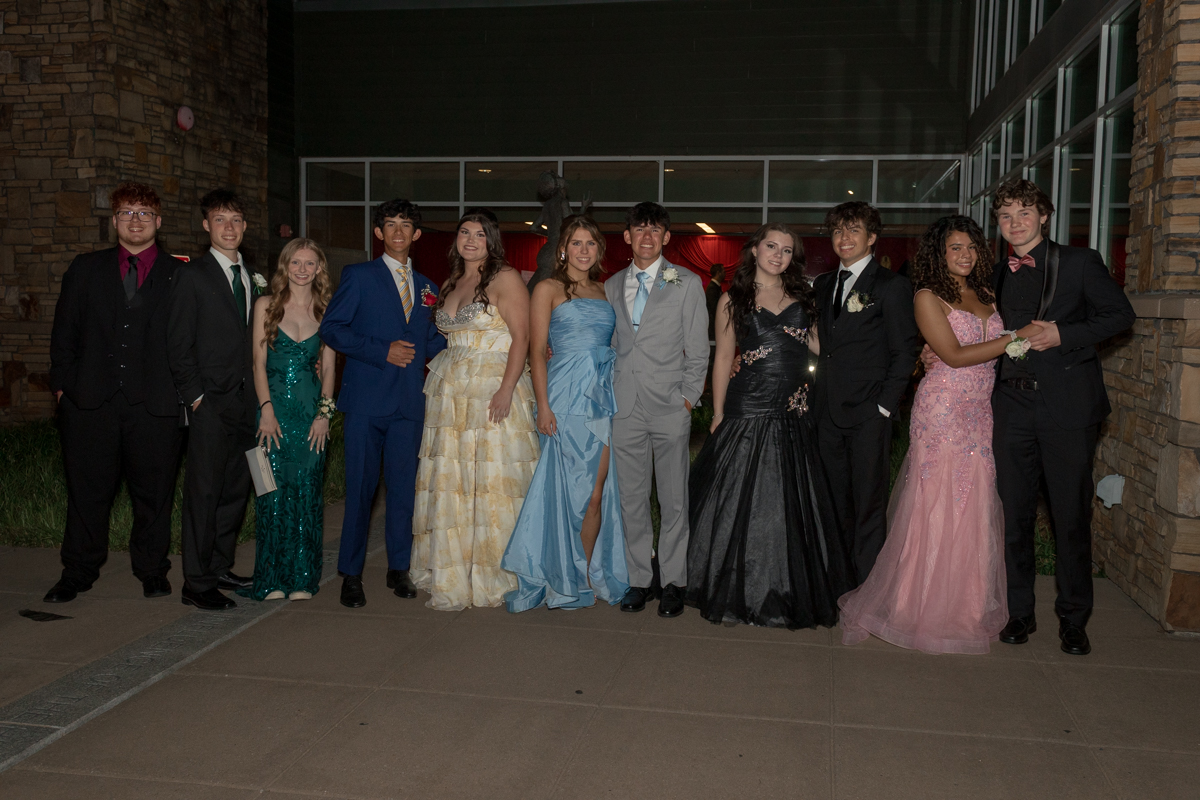 Group of teens in formal gowns and suits posing outside a building for a prom photo.