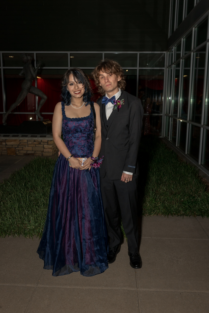 Young couple in formal attire posing outdoors at night; woman in a dark blue ball gown and pearls, man in a black suit with a blue bow tie and boutonniere.