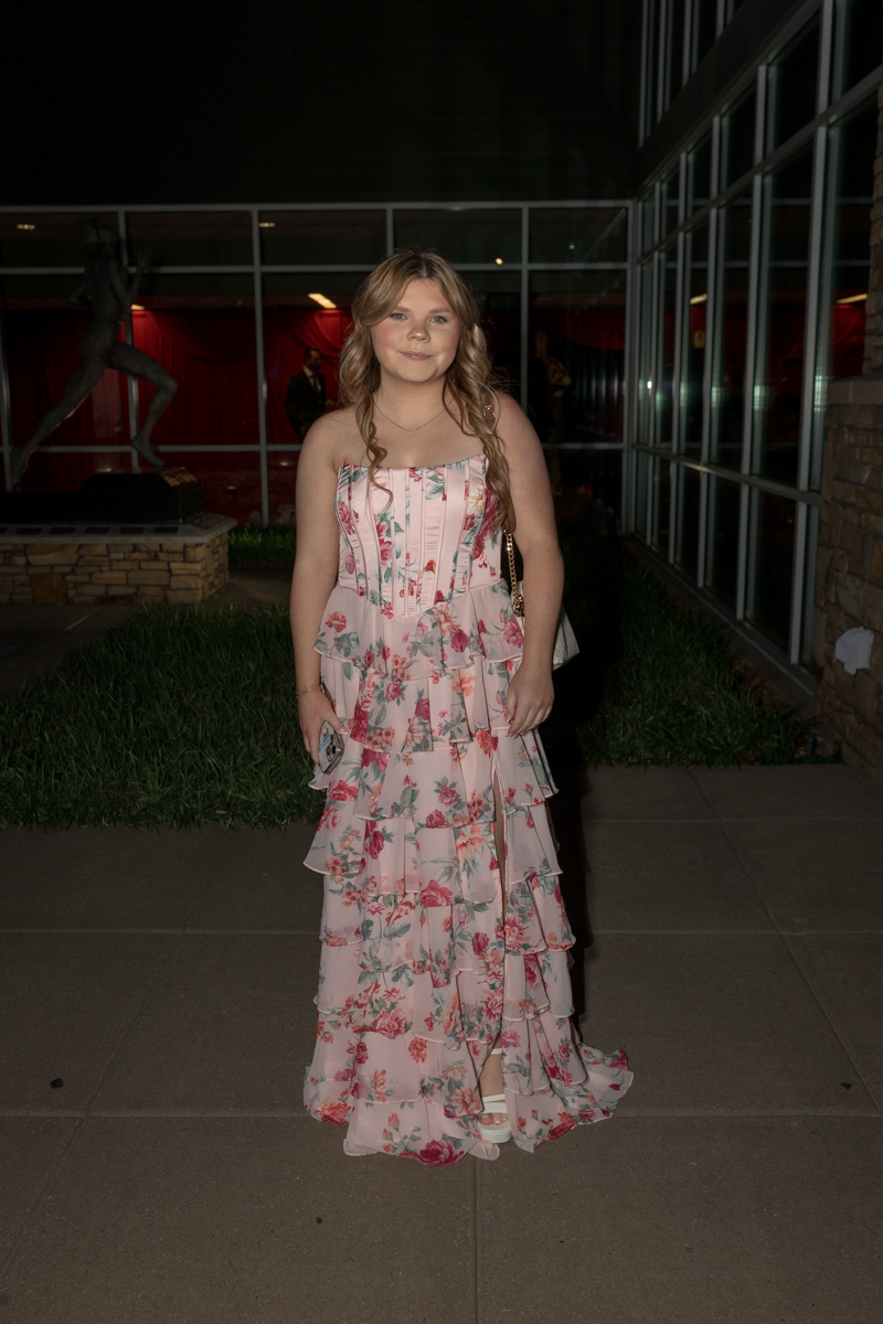 Young woman in a pink floral tiered dress standing outside a glass-walled building at night, holding a small clutch.