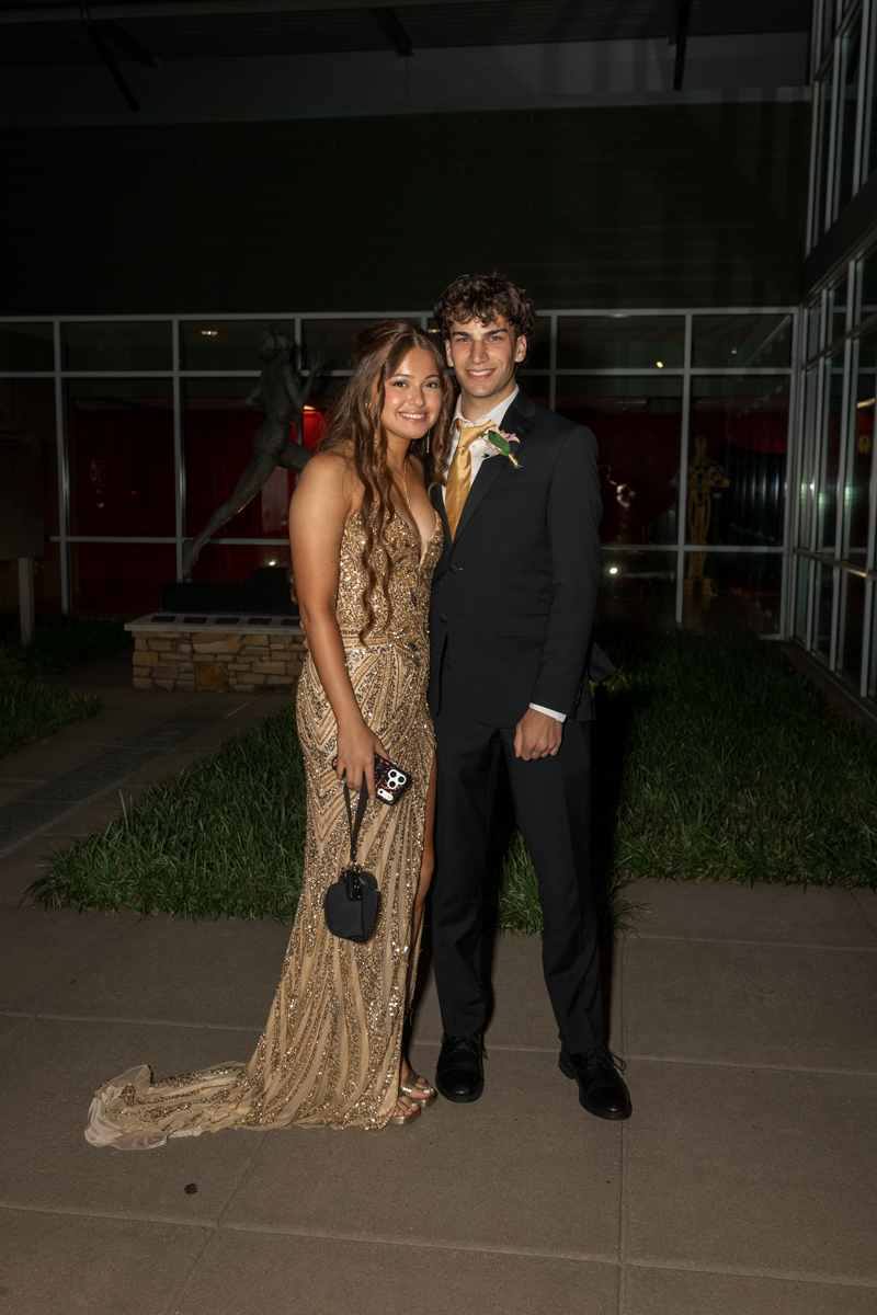Couple posing at a formal event: woman in a gold sequin gown with a small black handbag and a man in a dark suit with a gold tie and boutonniere, smiling at the camera outside a building at night.