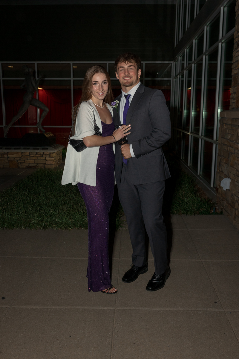 Couple dressed for a formal event posing together outdoors at night, the woman in a purple sequined gown and white shawl, the man in a gray suit and purple tie.
