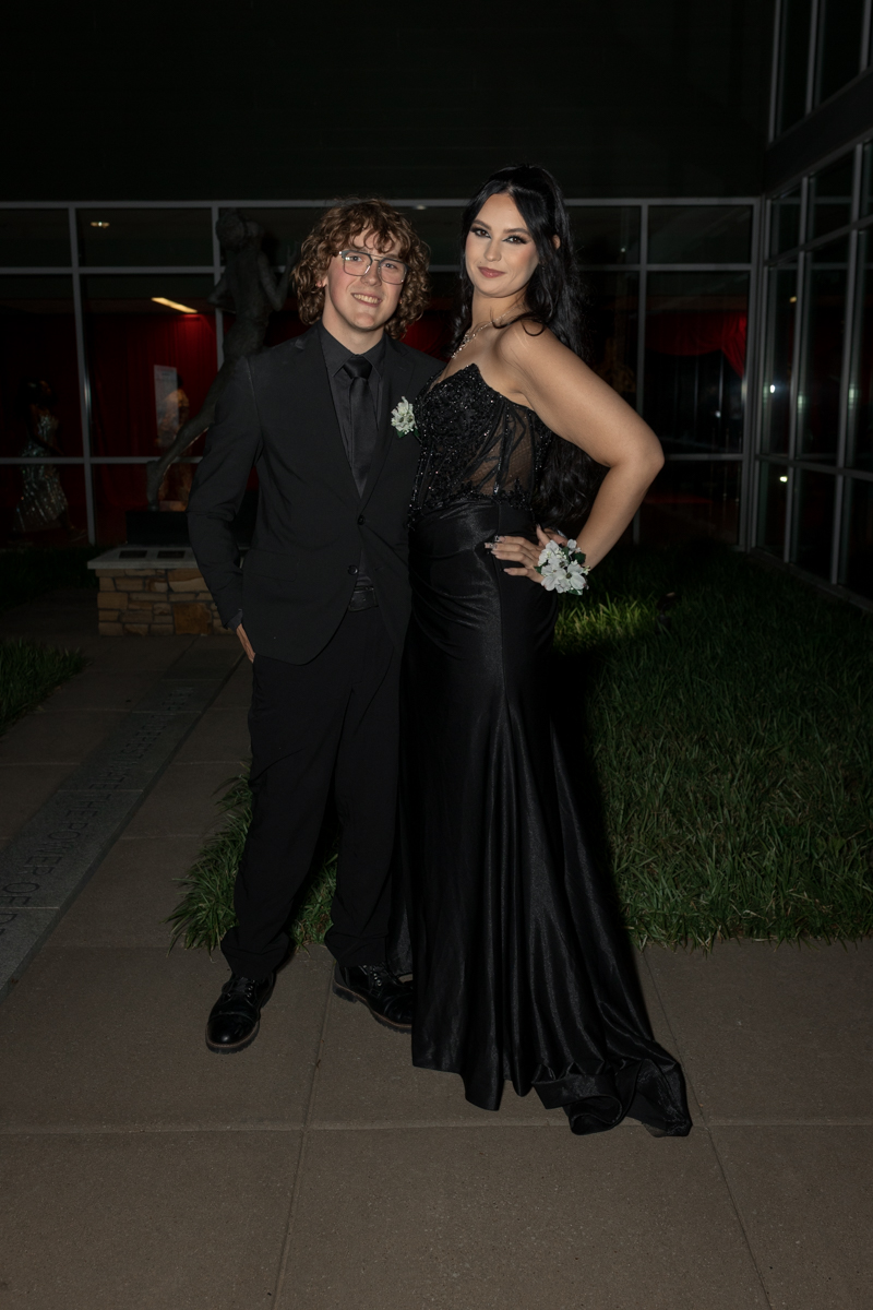 Two people in formal black attire pose together at night, smiling for the camera.
