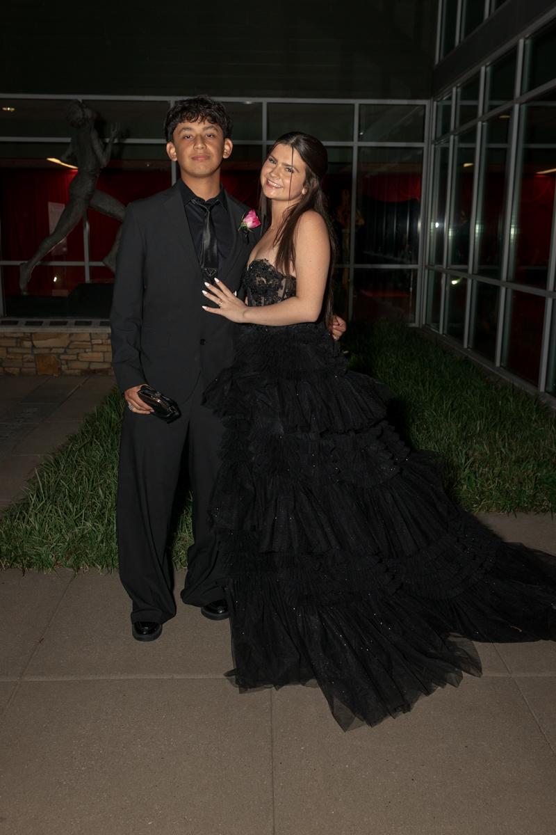 Two teenagers in formal attire posing together outside a glass-walled venue at night, smiling at the camera.