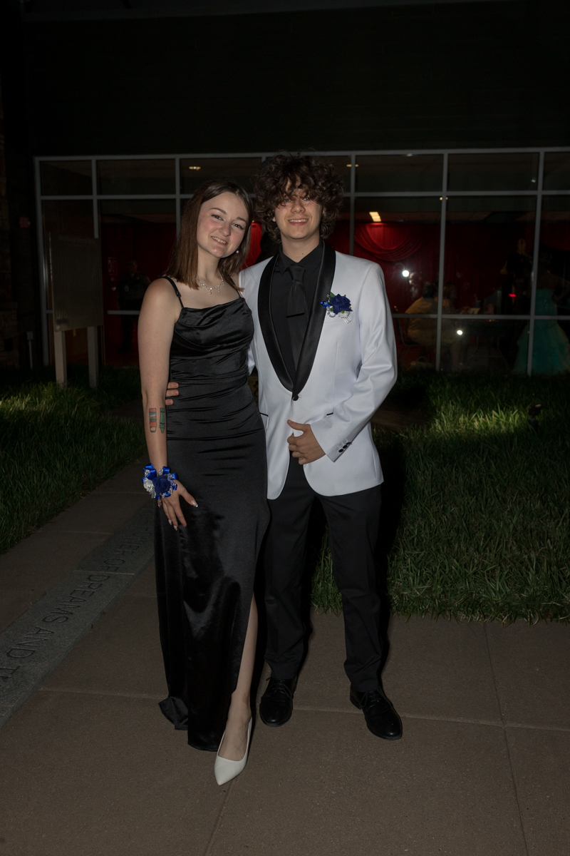 Two teenagers in formal wear pose outside at night: girl in a black satin gown with white heels and a corsage, and boy in a white tux with a black lapel and blue boutonniere.