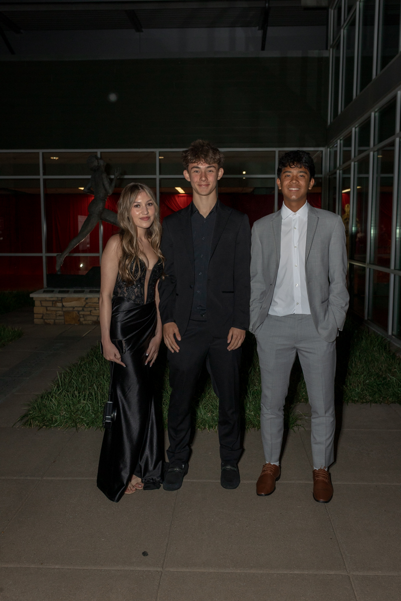 Three young adults in formal wear pose outside a venue at night: a woman in a black satin dress and two men in suits.