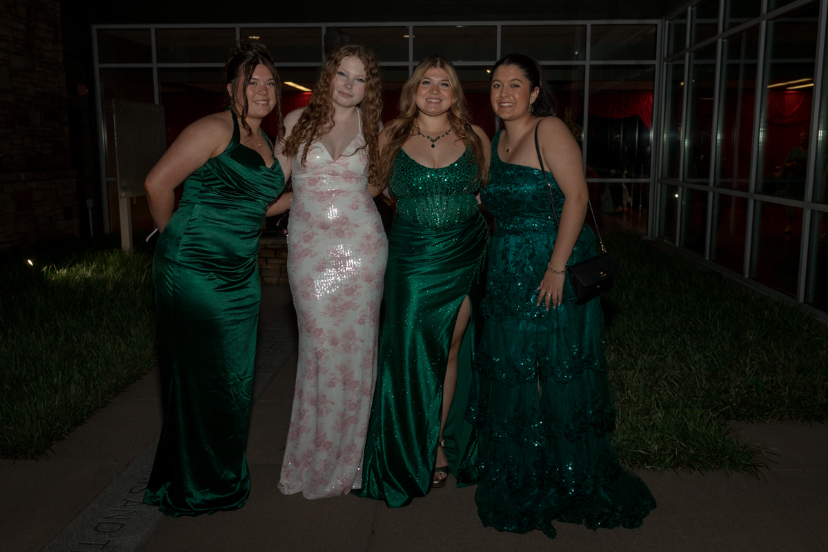 Four women in formal gowns pose together outdoors at night near a glass entrance to a building.