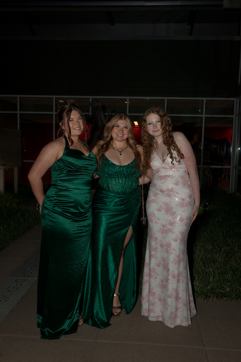Three women in formal gowns pose together outdoors at night, smiling for the camera.",