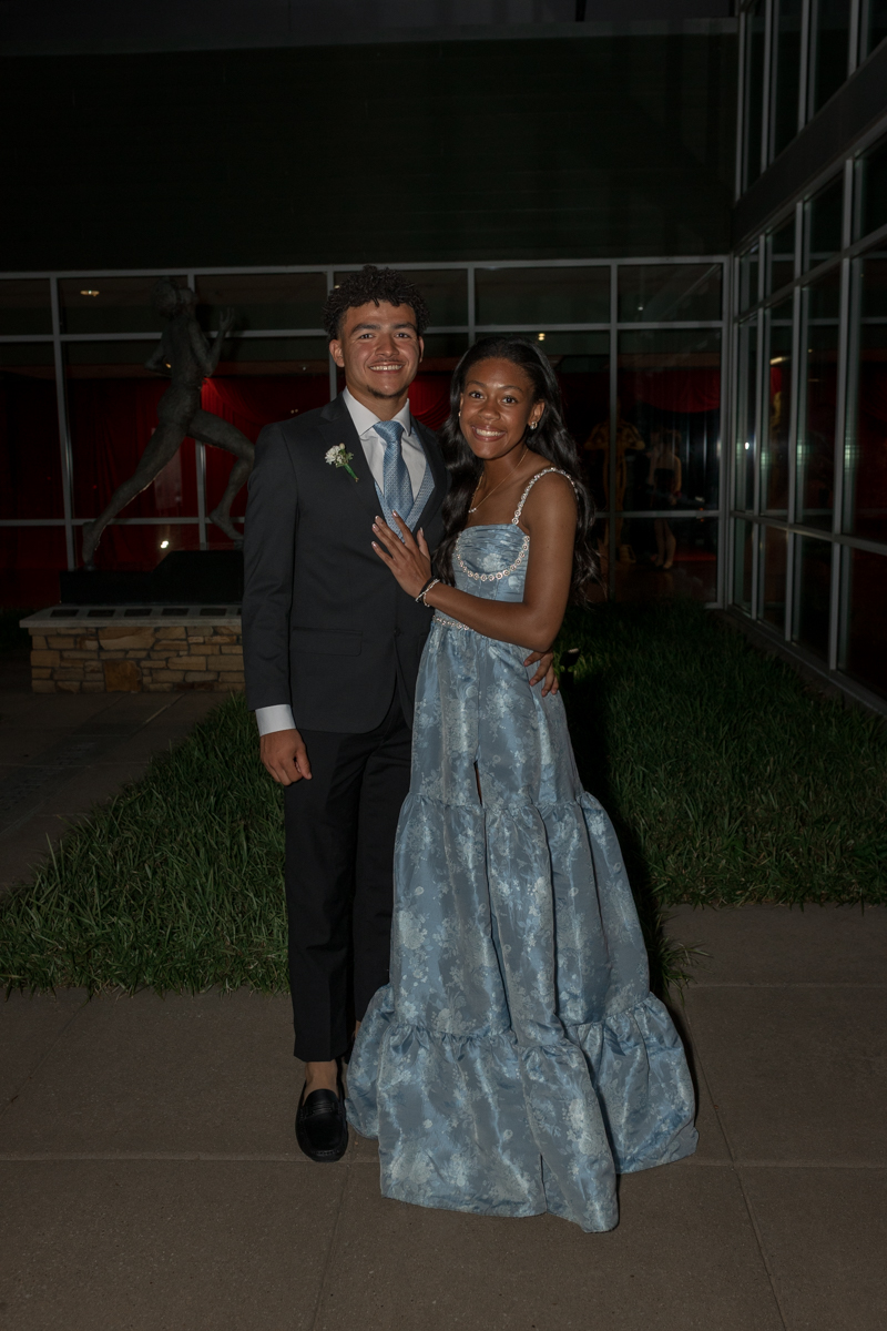 A smiling couple posing outside a building at night; the man in a suit and the woman in a pale blue formal gown."/A couple in formal attire posing together at an evening event.