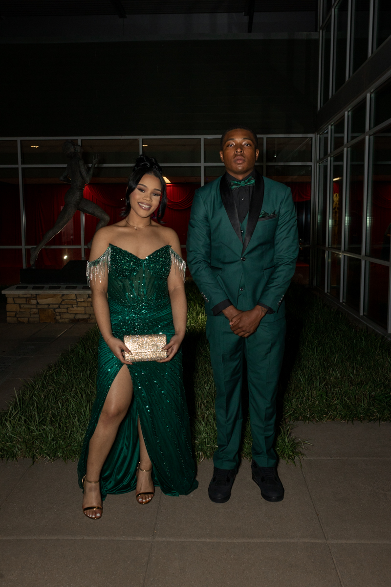 Couple in emerald formalwear posing outside a venue at night; woman in a sequined off-shoulder gown with a gold clutch, man in a green suit with black lapels and bow tie.