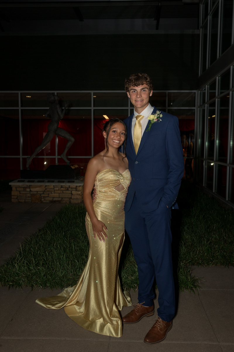 Two teenagers posing together at a formal event outside a glass-walled venue; girl in a sparkling gold gown and boy in a navy suit with a boutonniere, smiling at the camera (arms linked).