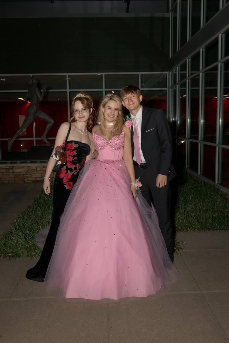 Three friends in formal attire pose together: center girl in a pink ball gown, left girl in a black dress with red floral embroidery, and a man in a suit with a pink tie on the right.