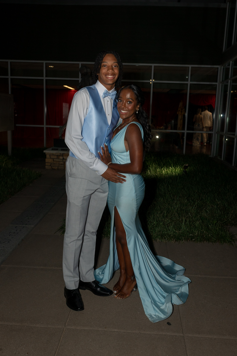 Smiling couple posing outdoors at night in formal wear: man in a light gray suit with a blue vest and bow tie, woman in a teal gown with a high slit.
