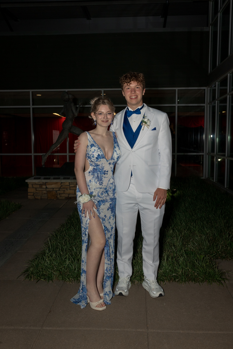 A young couple posing outside a building at night: the woman in a blue and white floral gown with a high slit, corsage on her wrist, and the man in a white suit with a blue vest and bow tie.