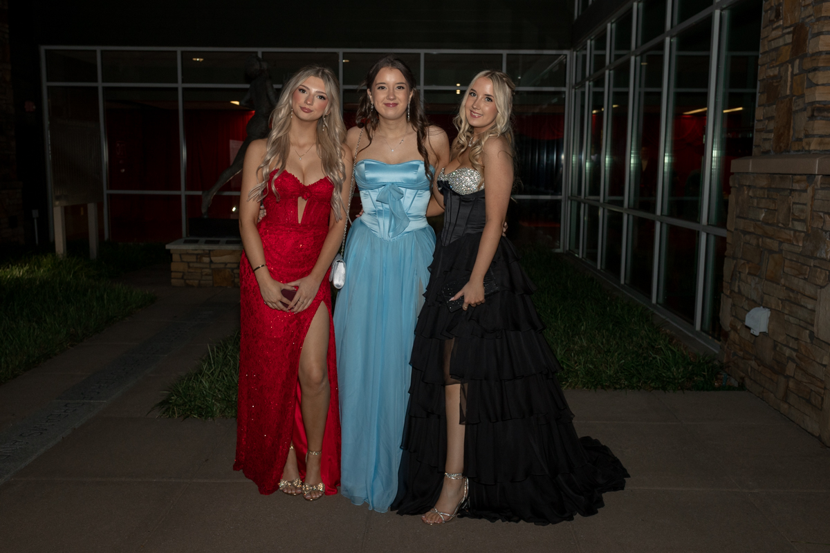Three young women in red, blue, and black formal gowns pose together outside a building at night, smiling for the camera (event photo).