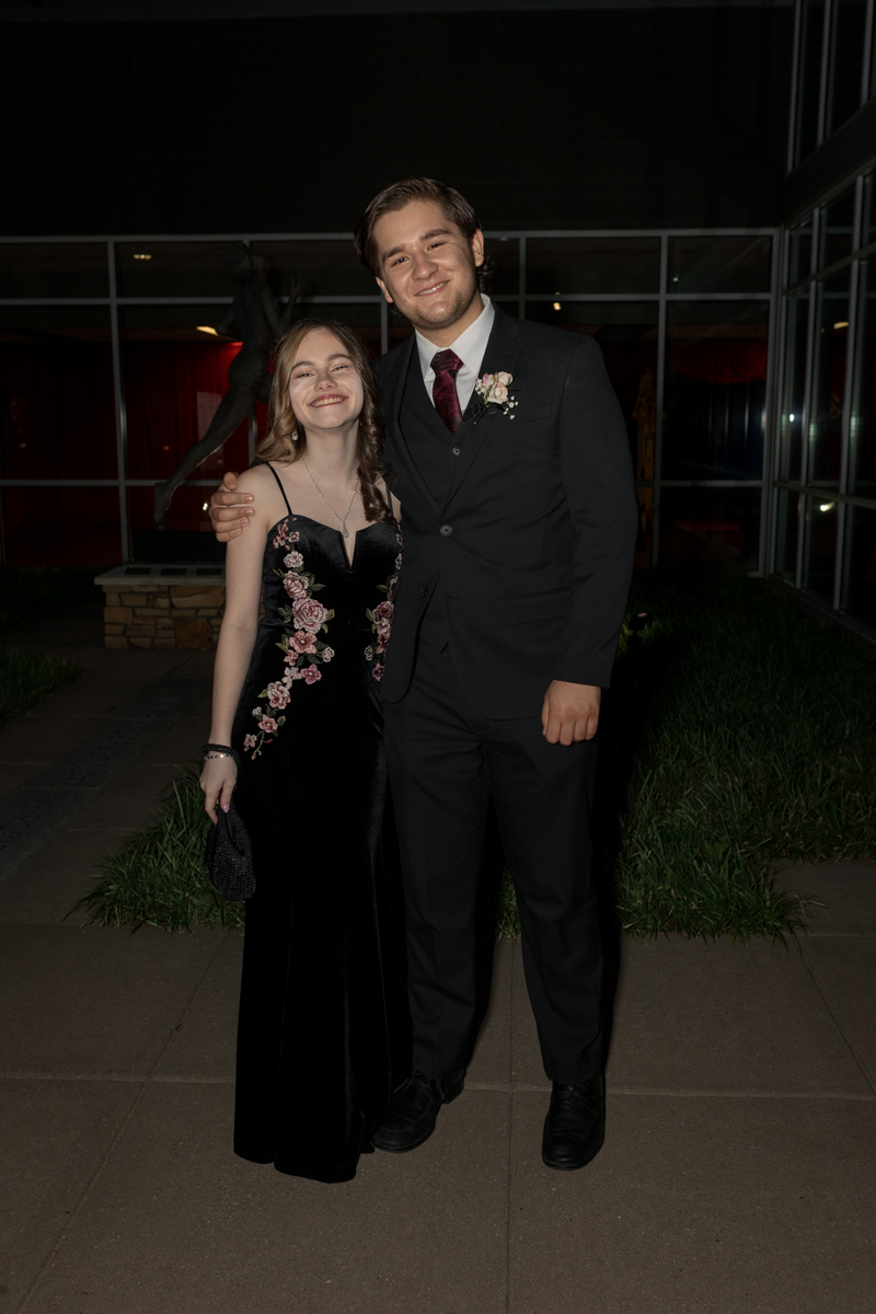 A young couple dressed formally stands arm in arm, smiling for the camera outside a glass-walled building at night; the woman wears a black velvet floral dress, the man a black suit with a boutonniere.