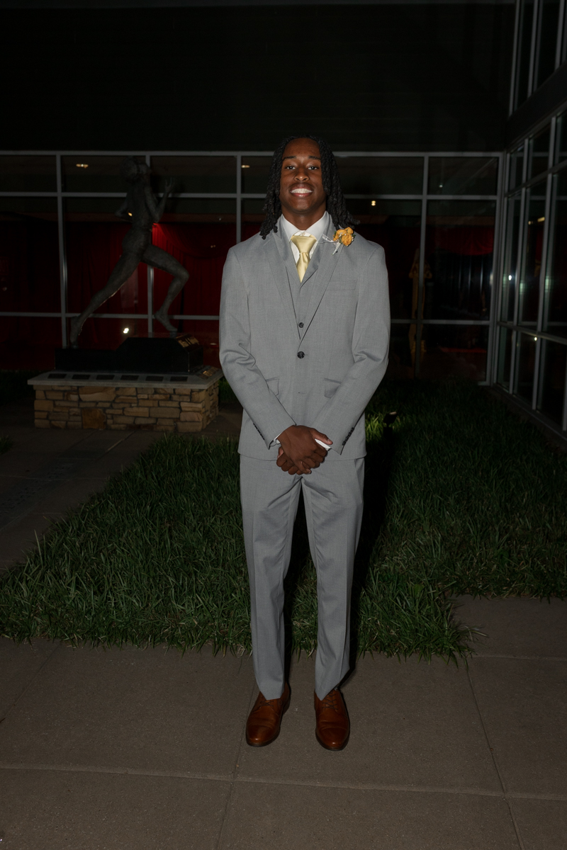 Young man in a light gray suit with a yellow tie and boutonniere, standing outside on a nighttime event, hands clasped.