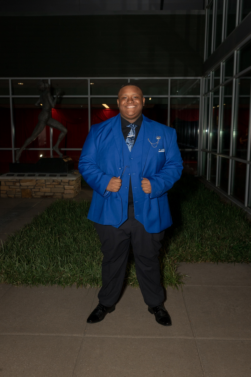 Smiling man in a bright blue blazer and vest posing with hands on his blazer outside a glass building at night.