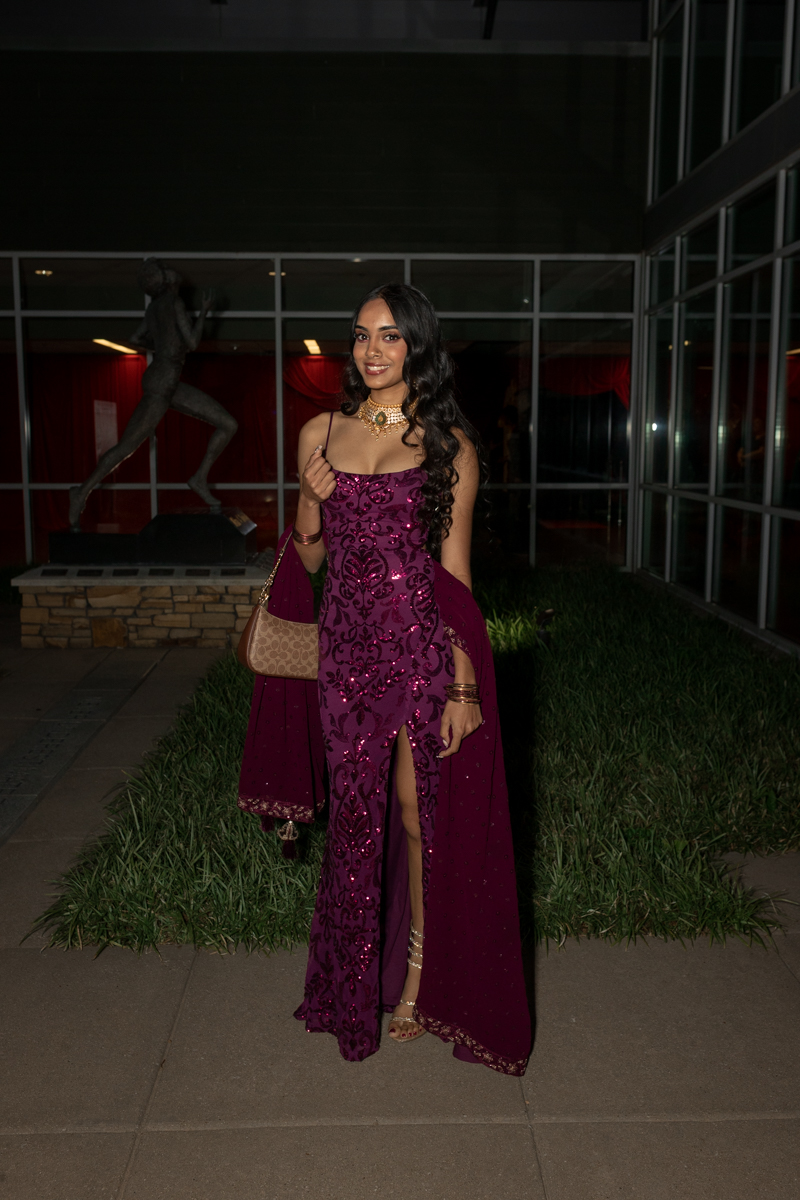 Woman in a magenta sequined gown with a thigh-high slit, posing outside at night with a beige purse and gold jewelry.