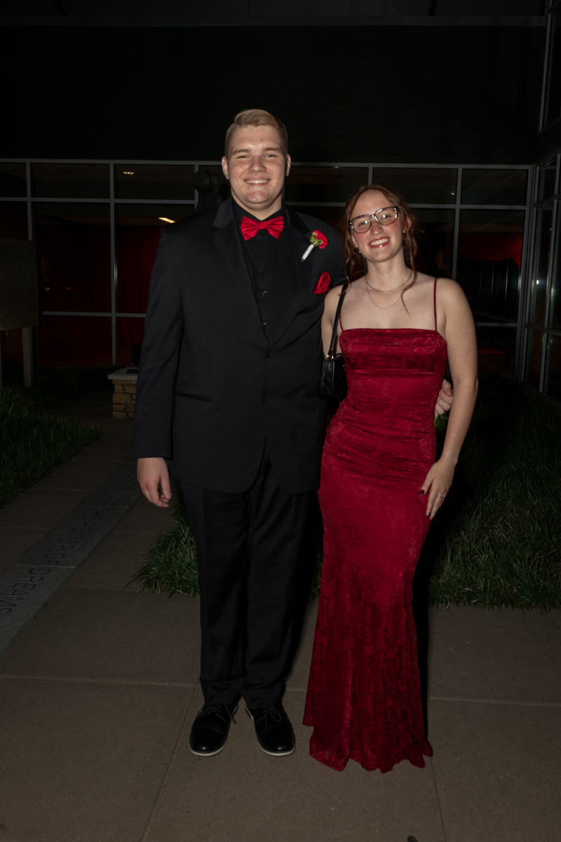 Two young adults in formal attire posing together: man in black tux with red bow tie and boutonniere, woman in a red velvet gown with glasses.