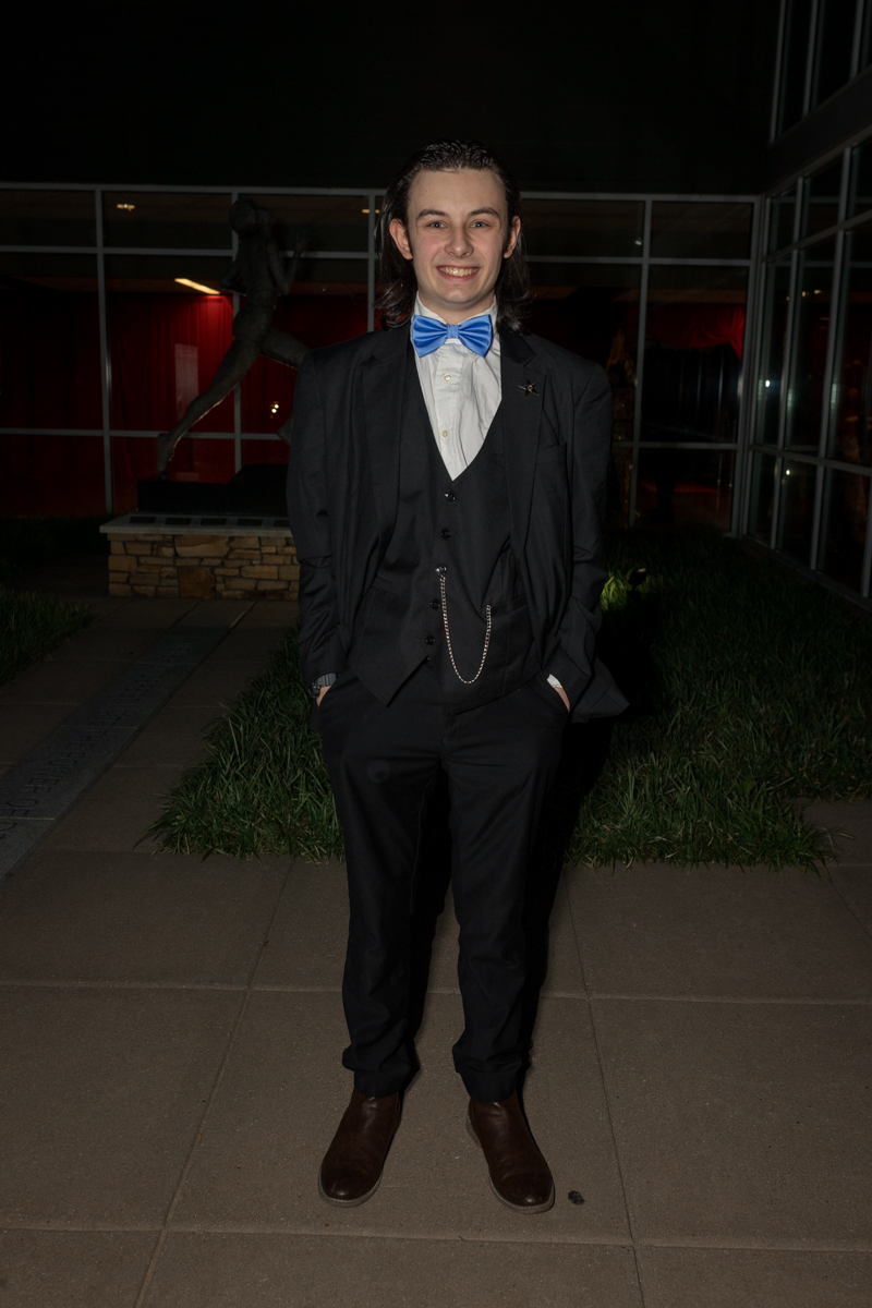 Young man in a black suit with a blue bow tie, vest, and pocket chain, standing outdoors at night and smiling.