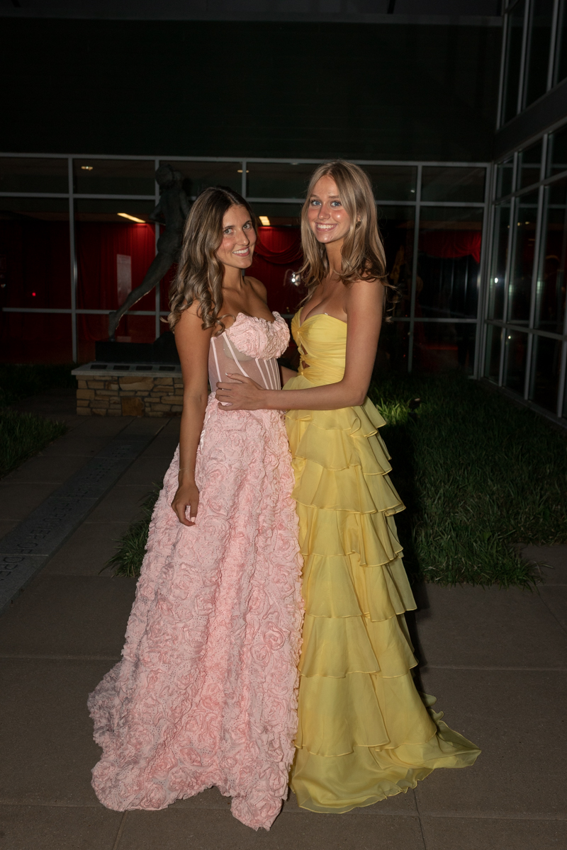 Two young women in formal gowns pose together outside a modern building at night; one in a pink strapless floral dress, the other in a yellow tiered dress, both smiling.