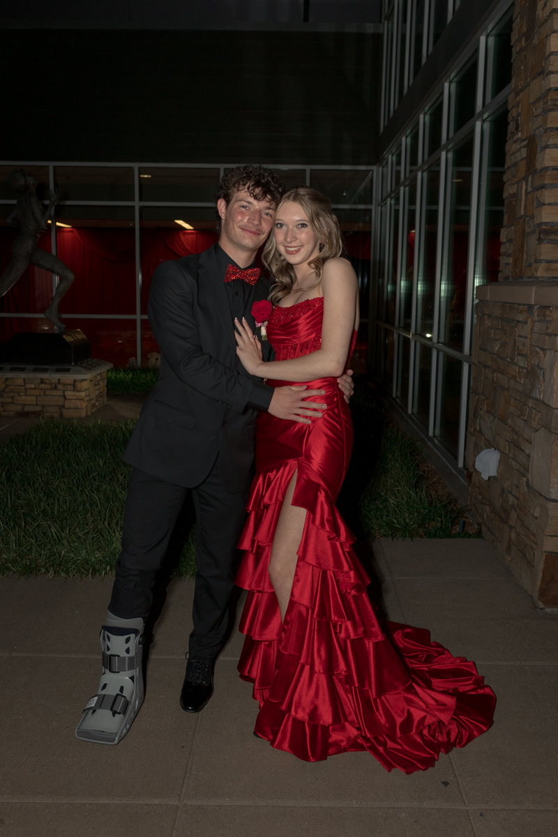 A young couple in formal attire posing outside a glass-front building; the man wears a black suit with a red bow tie and a protective medical boot on his left foot, while the woman wears a red strapless gown with a high slit and ruffled layers, and they smile at the camera.