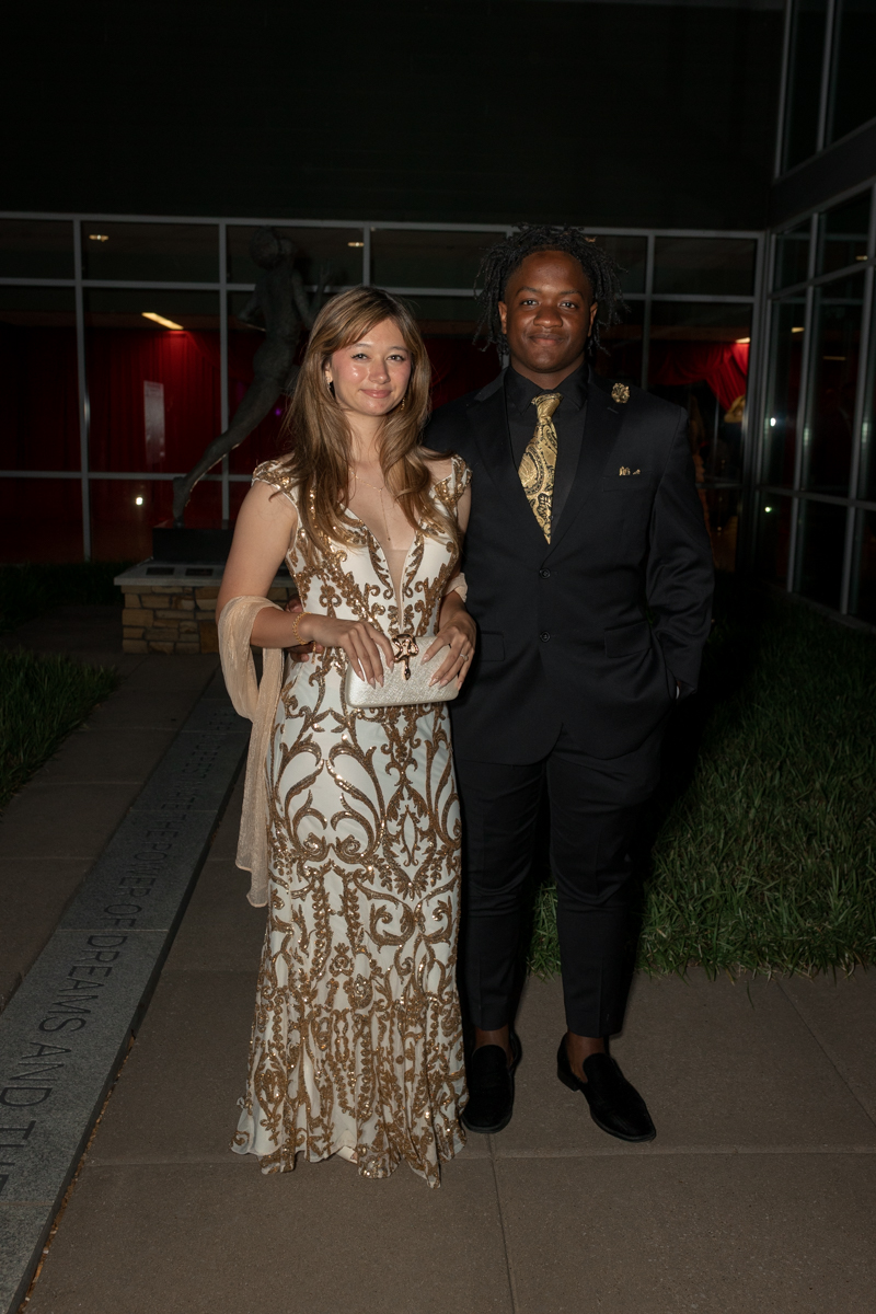 A young couple posing outdoors at night: woman in a cream and gold embellished gown with a sheer shawl, standing beside a man in a black suit and gold tie.
