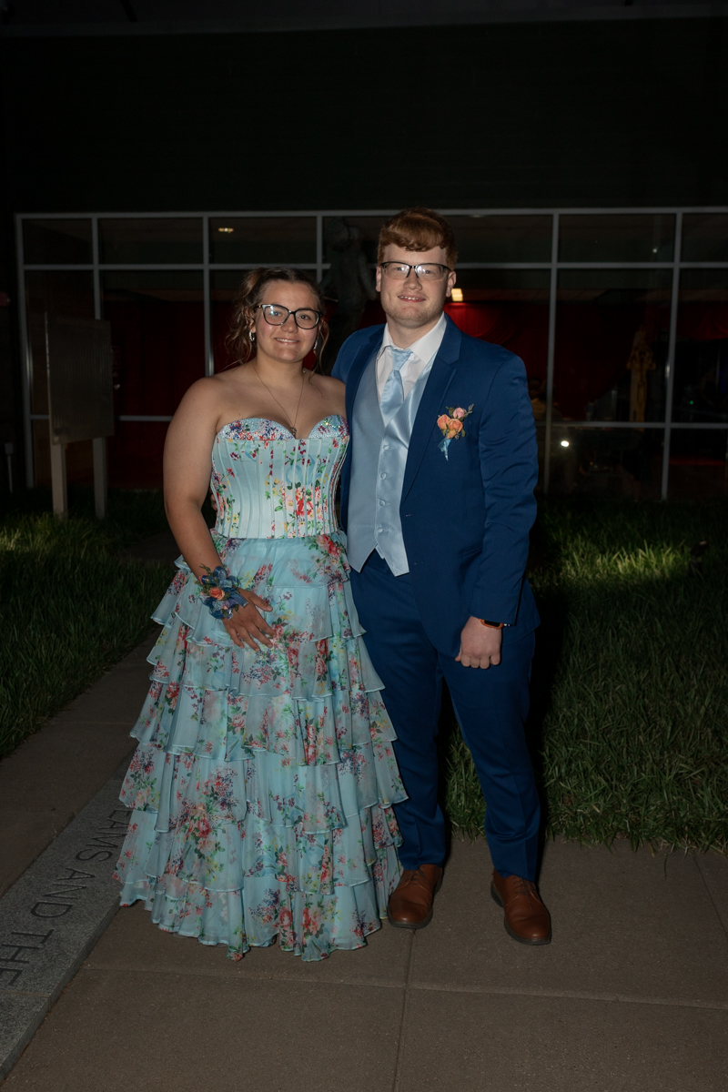 Two young adults pose outdoors at night: a woman in a light blue floral strapless ballgown with ruffled layers and a corsage, and a man in a blue suit with a matching light blue vest and tie, standing on a sidewalk by grass.
