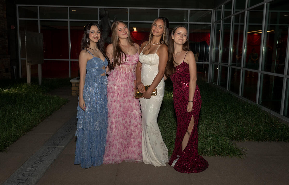 Four young women in formal gowns posing outdoors at night, smiling for a group photo in blue, pink, white, and burgundy dresses.