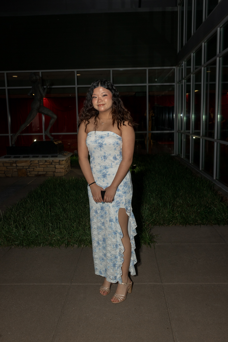 Young woman in a strapless blue floral dress with a high slit, standing outdoors at night near glass windows and a lawn.