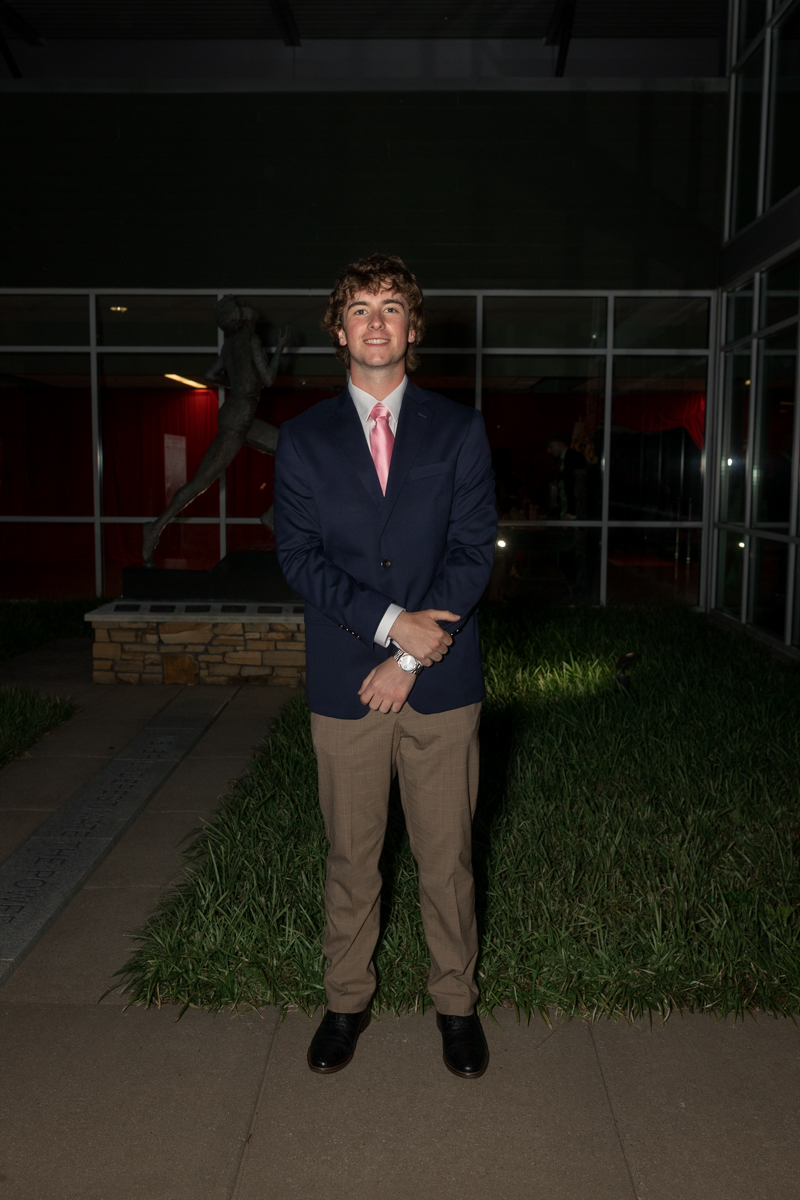 Young man in a navy blazer, pink tie, and khaki pants posing outdoors at night on a sidewalk.