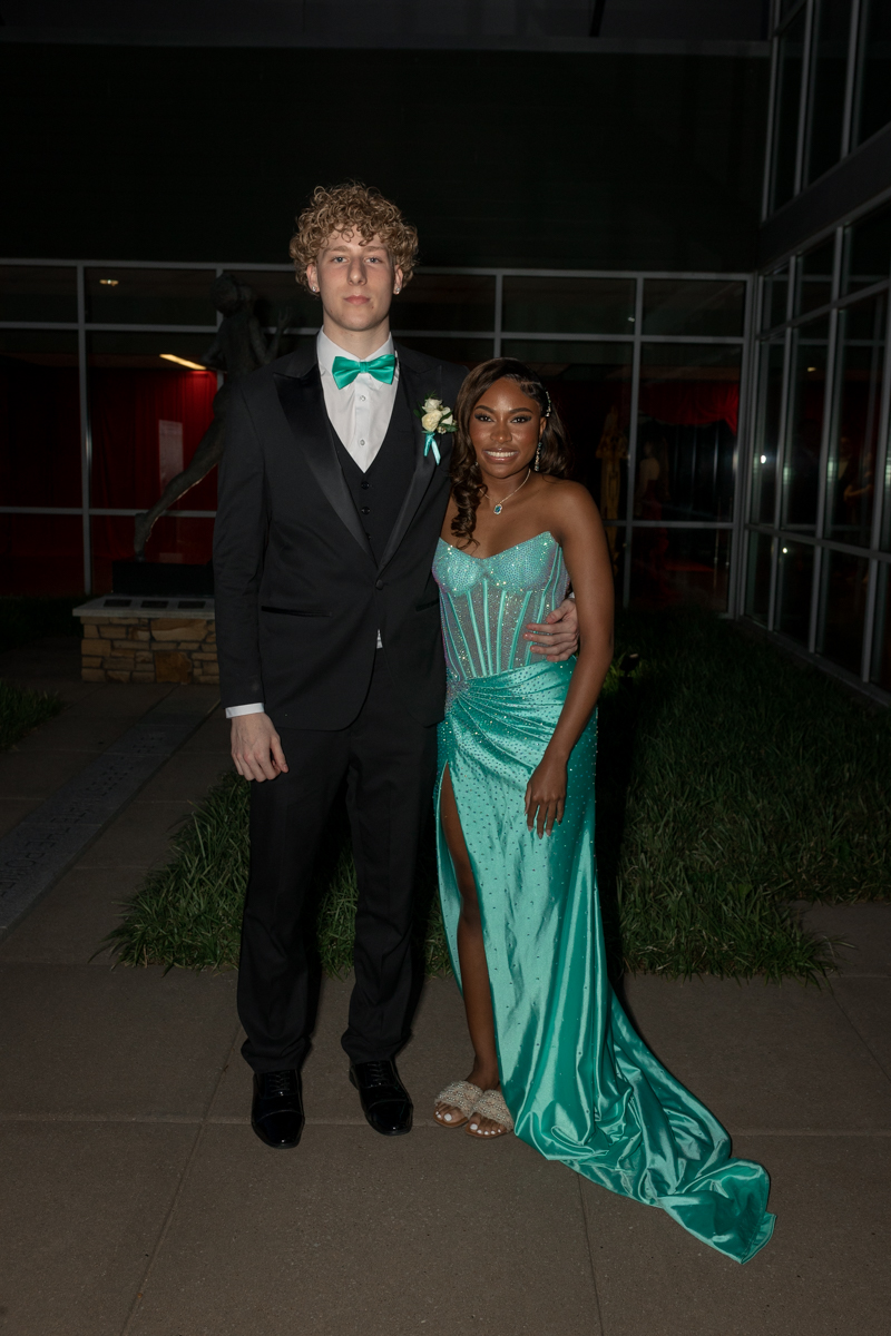 Couple dressed for a formal event posing outdoors, the man in a black suit with teal bow tie and boutonniere, the woman in a turquoise strapless gown with a high slit and silver jewelry.