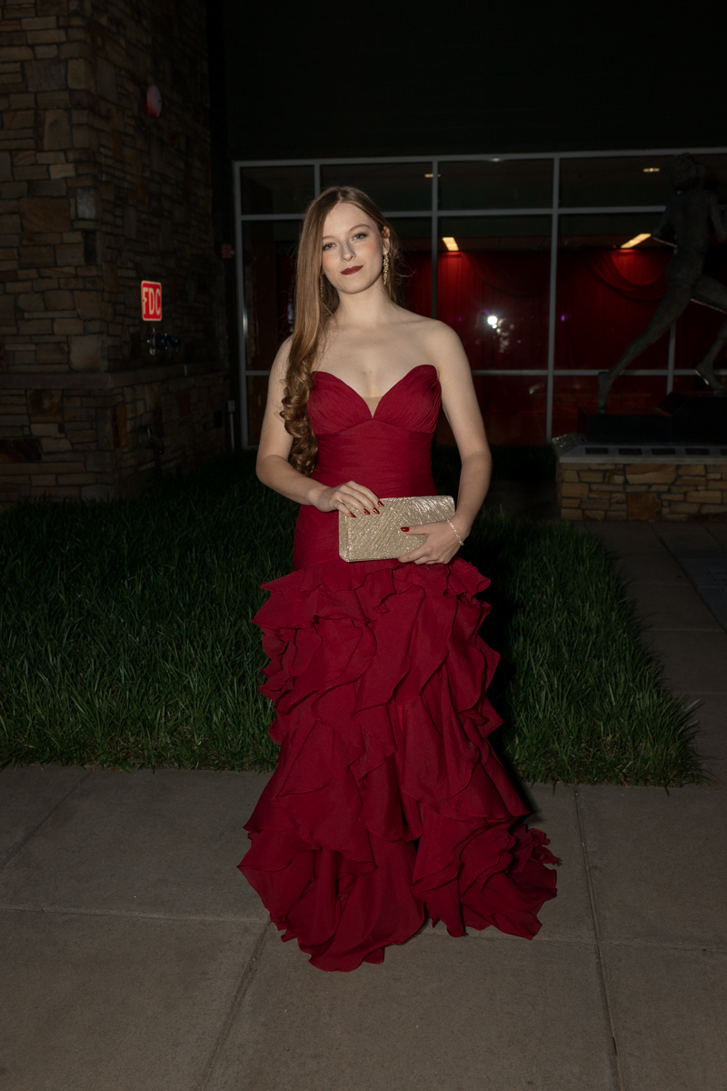 Woman in a strapless red gown with a ruffled skirt holding a gold clutch outside a building at night