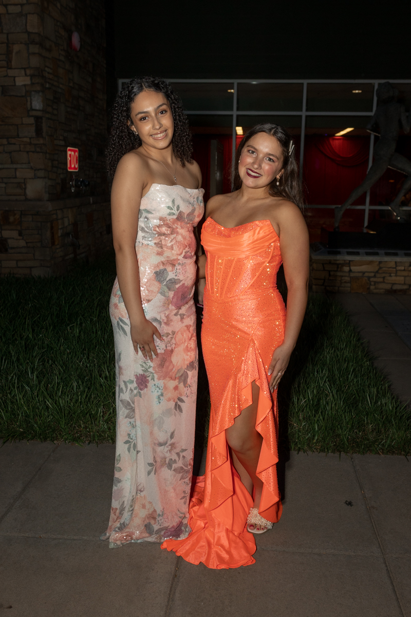 Two young women in formal gowns posing outdoors at night; one in a pink floral strapless dress, the other in a bright orange sequin gown.