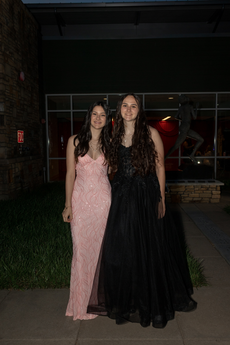 Two young women in formal gowns standing side by side outside a building at night, one in a pink sparkly dress and the other in a black gown, smiling down at the camera.