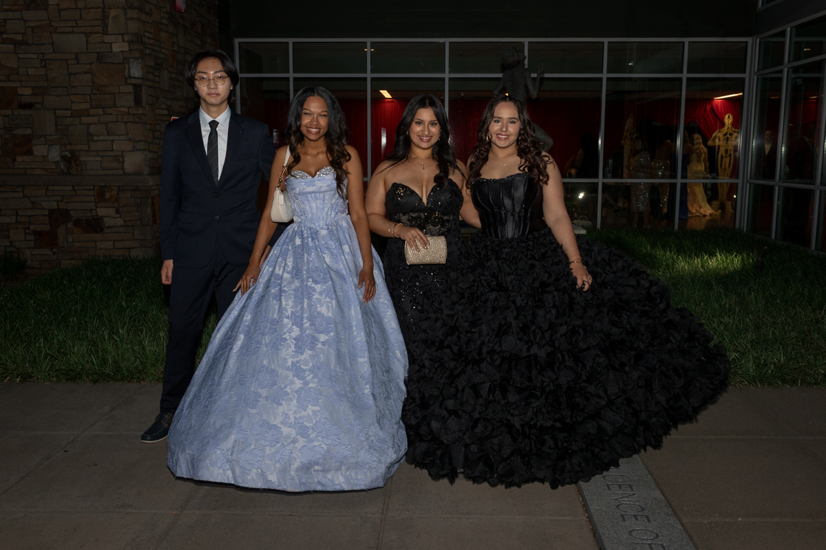 Four friends pose in formal wear outdoors: a man in a dark suit and tie with three women in ball gowns (blue lace, black sequined, and black ruffled) in front of a glass building.