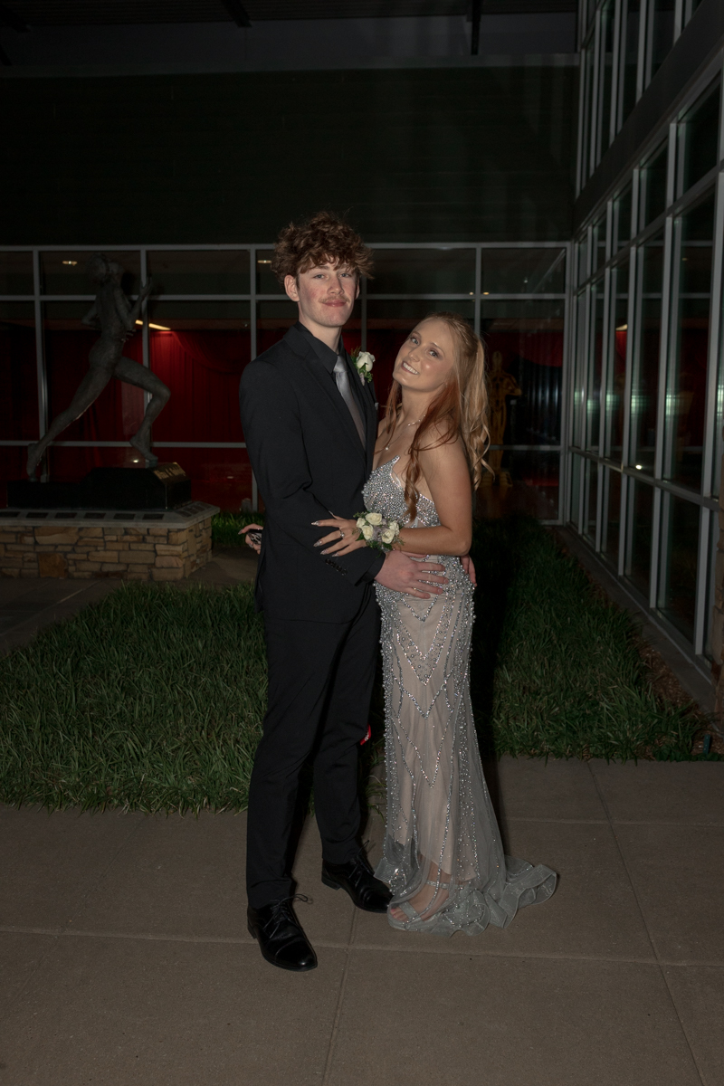 Young couple in formal wear posing together outside a glass-walled venue at night.