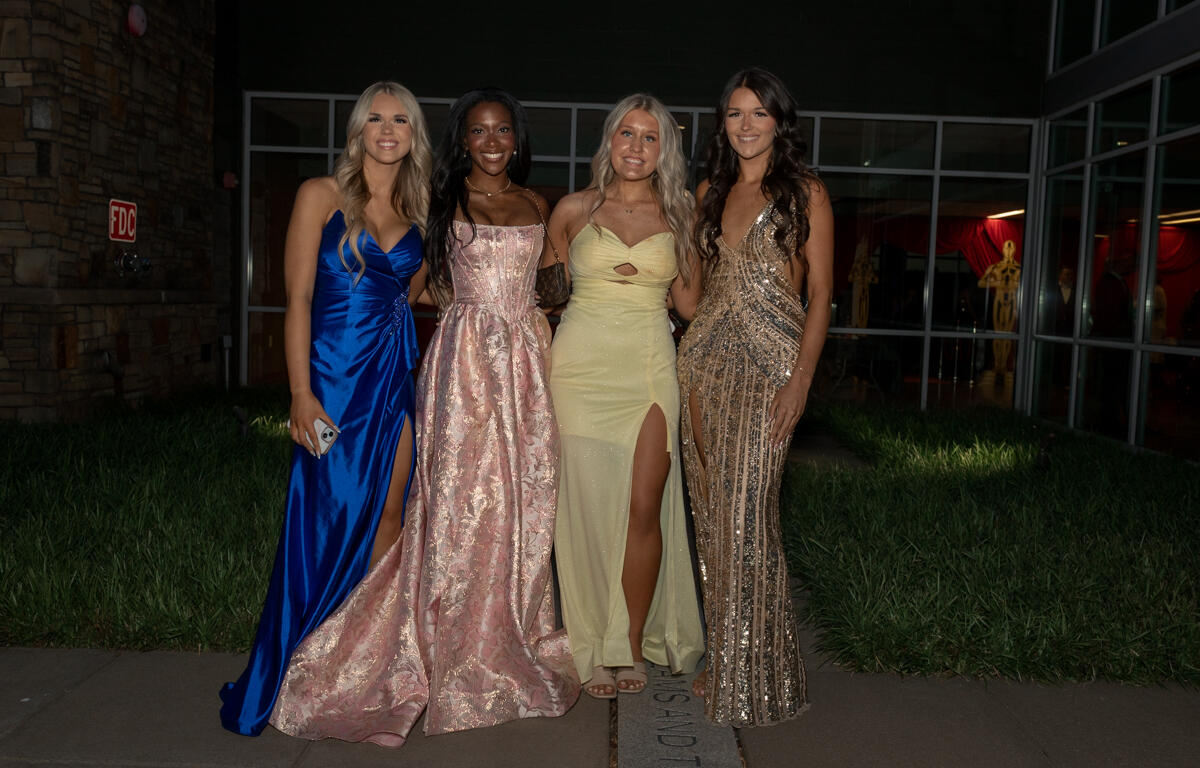 Four women in formal gowns pose together outside a building at night, smiling for the camera during a formal event.