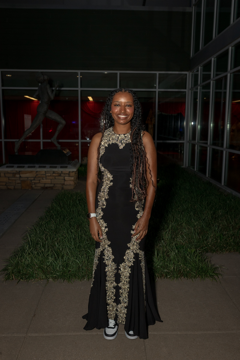 Woman in a floor-length black gown with gold embroidered details standing outdoors at night, smiling at the camera.
