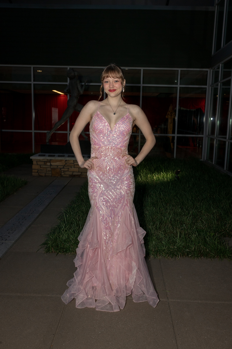 Young woman in a pink sequined evening gown posing with hands on hips outside a glass-building backdrop at night.