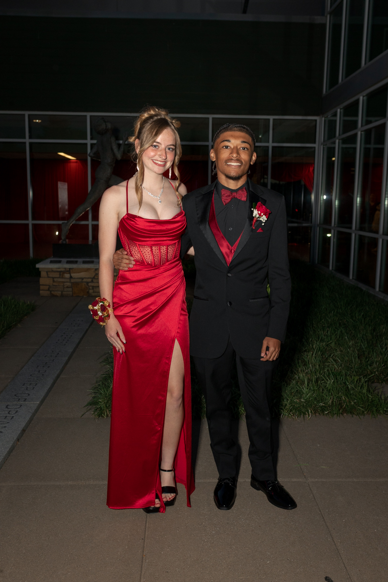Couple in formal wear posing together at night; woman in a red satin gown with a thigh-high slit, man in a black tuxedo with a red bow tie and vest.